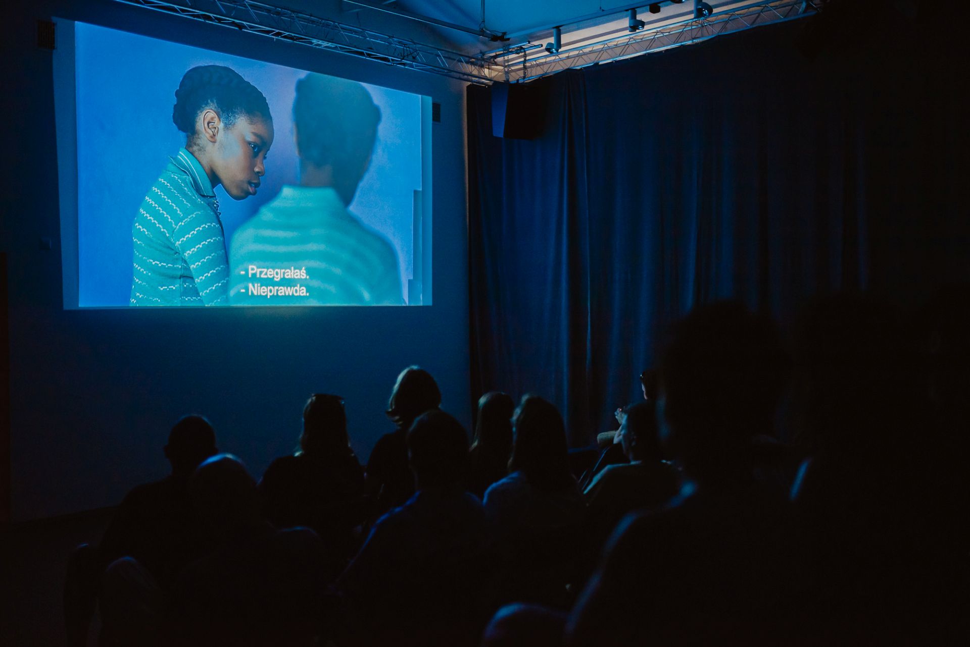Ludzie siedzą na krzesłach w sali widowiskowej i oglądają film wyświetlany na ścianie.