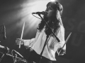 Ponownie zdjęcie z koncertu Asea Sool. Widzimy wokalistkę śpiewającą do mikrofonu oraz stojącą przed perkusją. Zdjęcie czarno- białe.
