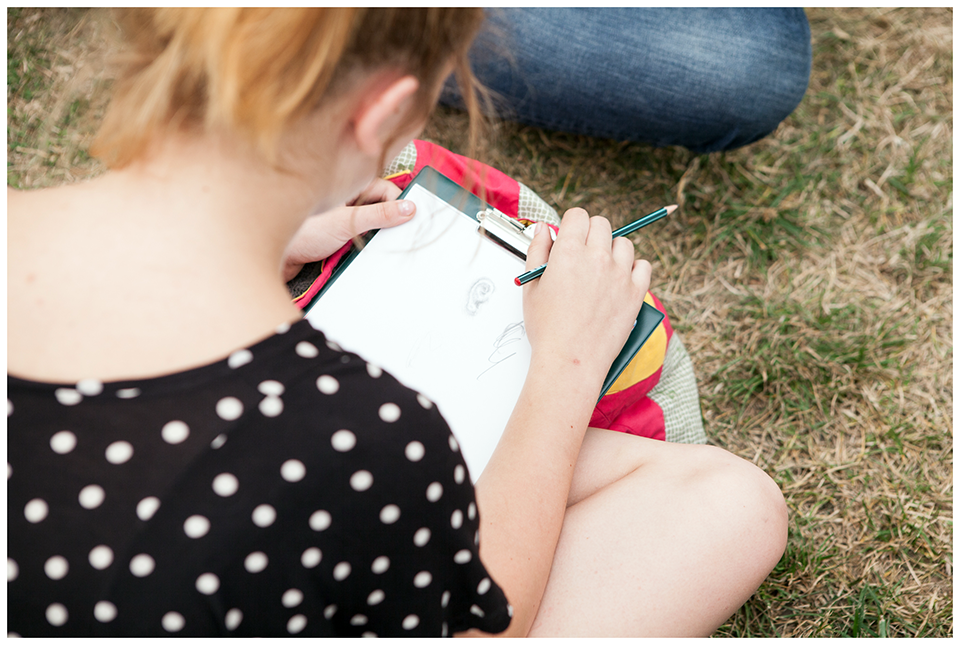 [Zdjęcie zrobione zza ramienia siedzącej na trawie dziewczyny. Młoda kobieta szkicuje ołówkiem na kartce papieru.]