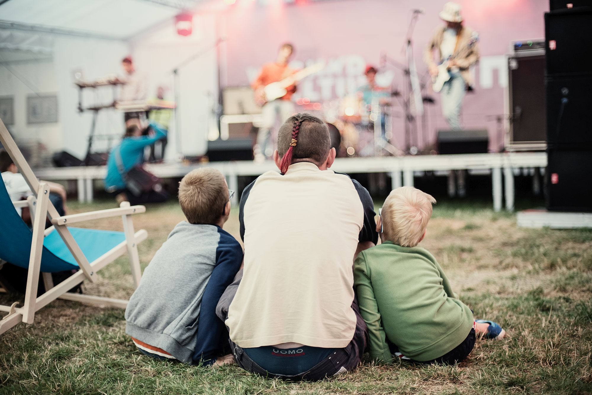 [Mężczyzna z wraz z dwójką dzieci po jego obu stronach siedzą tyłem do obiektywu na trawie, pod namiotem festiwalowym. Zwróceni są w kierunku występującego na scenie zespołu.]