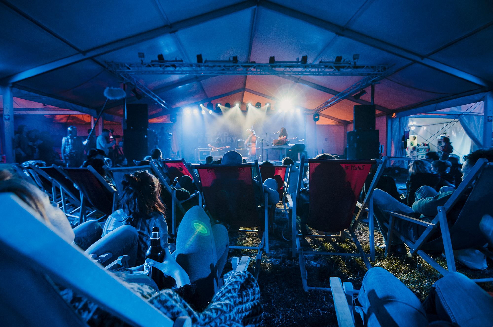 [Zdjęcie z perspektywy publiczności. Widzowie leżą na leżakach. Na dalszym planie zespół gra na scenie. Wszyscy oświetleni są niebieskim światłem.]