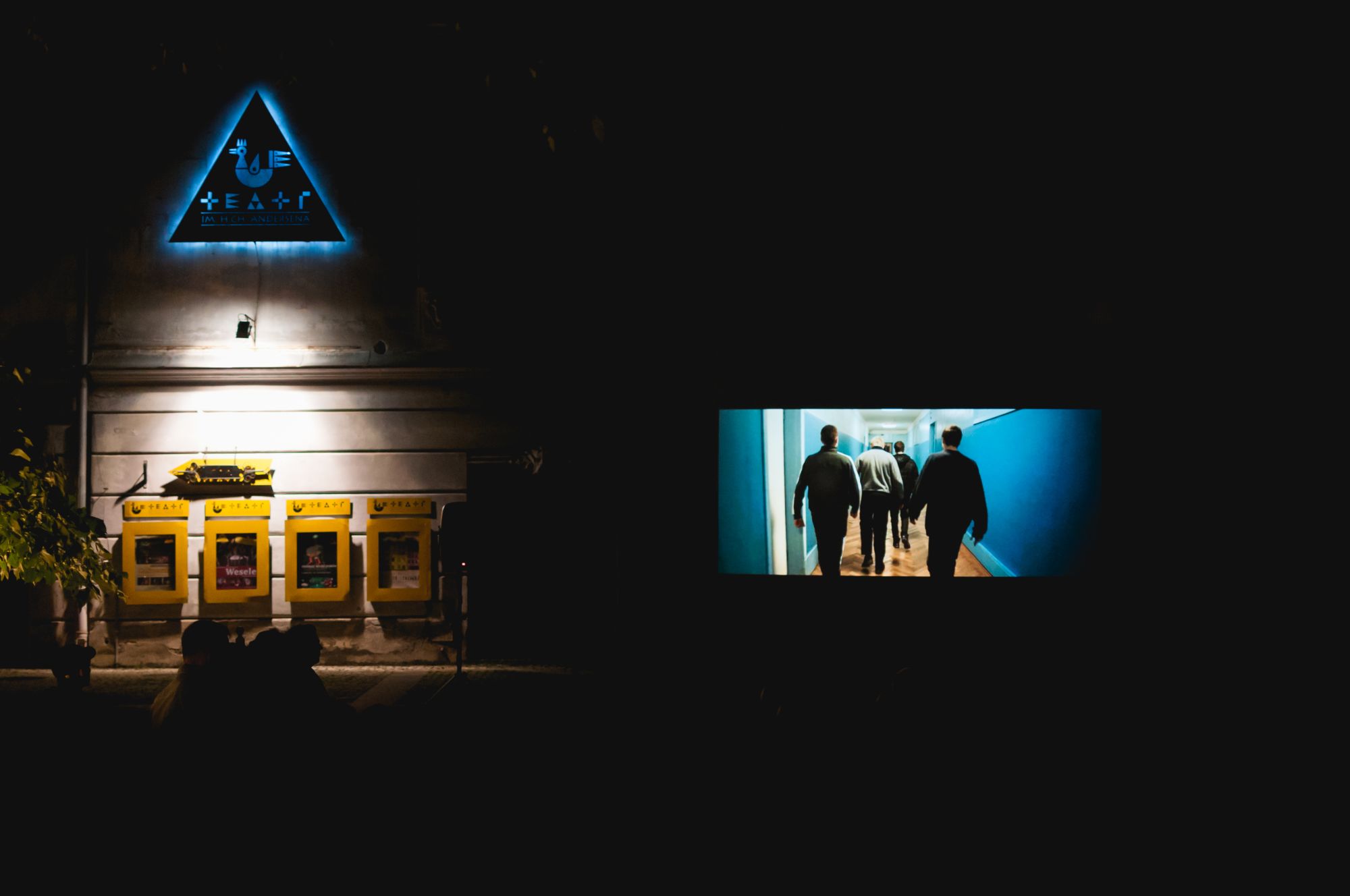 [Plac przed Teatrem Andersena. Wieczorny seans filmowy w ramach wydarzenia Move East Movie. Z lewej strony widoczne logo teatru, z prawej ekran z wyświetlanym filmem.]