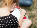 [Zdjęcie zrobione zza ramienia siedzącej na trawie dziewczyny. Młoda kobieta szkicuje ołówkiem na kartce papieru.]