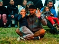 [Klub festiwalowy. Mężczyzna siedzi po turecku na trawie z dzieckiem. Obydwoje machają w stronę obiektywu. Na drugim planie uczestnicy festiwalu siedzący na leżakach.]