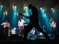 [Zdjęcie z koncertu zespołu Hańba przedstawia ciemną sylwetkę mężczyzny grającego na gitarze. W tle trzech mężczyzn w beretach, białych koszulach i kolorowych spodniach grających na instrumentach muzycznych.]