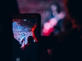 Na zdjęciu widać rejestrowany koncert przy użyciu telefonu komórkowego, na ekranie którego widać muzyka. W tle rozmazana postać artysty grającego na scenie.