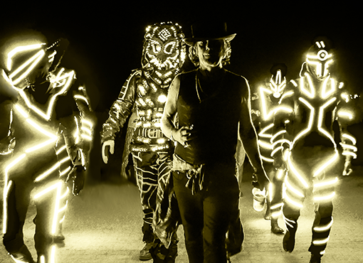 Juno Reactor & Mutant Theatre podczas występu. W centrum mężczyzna ubrany na czarno w kapeluszu. Dookoła niego postaci w świecącyck kostiumach, z maskami tygrysów na twarzach.
