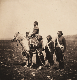 Zdjęcie czarno-białe, z dawnych lat. Czarnoskóry mężczyzna na koniu. Dookoła niego troje innych mężczyzn.