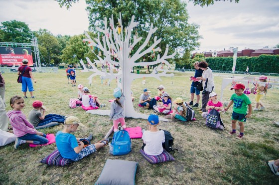 Dzieci siedzą na poduszkach na trawie, dookoła drzewa z kartonu. Wieszają na nim zrobione przez siebie ozdoby.