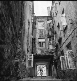 Czarno-Biała fotografia Starego Miasta. Widzimy wąską uliczkę i kamienice. Okna otwarte są na zewnątrz.