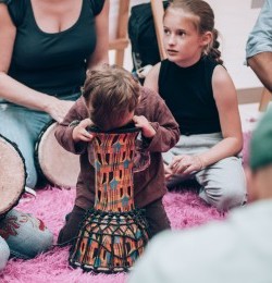 Dzieci siedzą na różowej wykładzinie. Uczą się grać na djembe.