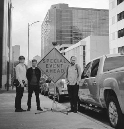 Czarno-biała fotografia członków zespołu "Trupa Trupa". Muzycy stoją na ulicy obok auta i znaku drogowego "Special Event Ahead".