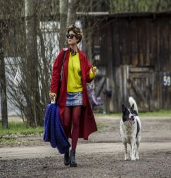 Stopklatka z filmu "Kryształ" w reżyserii Darii Żuk. Przedstawia kobietę ubraną bardzo kolorowo, która idzie przez wieś z psem. W tle widać drewniane zabudowania.