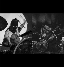 Fotografia czarno-biała. Członkowie zespołu BNNT grający koncert na scenie. Perkusista ma na twarzy kominiarkę.