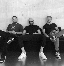 Fotografia czarno-biała. Członkowie zespołu Trio siedzą na kanapie pod ścianą.