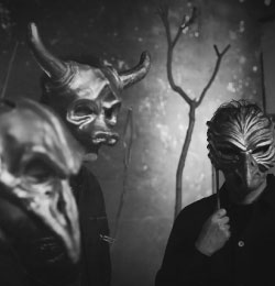 Fotografia czarno-biała. Trzy osoby z maskami na twarzach. Maski są podobiznami zwierząt: byka i ptaków.