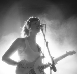 Czarno-biała fotografia Lucy Kruger i The Lost Boys. Kobieta gra na gitarze i śpiewa do mikrofonu.